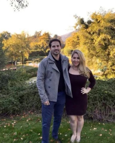Couple standing in grassy garden with hedges, trees, and mountains in background