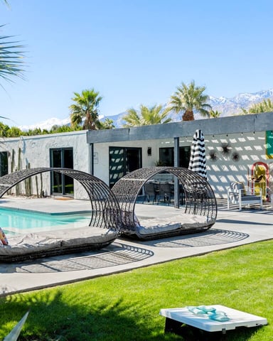Modern poolside patio with turquoise pool, striped umbrella, woven lounge pods, white building, palm trees, and snow-capped mountains in background