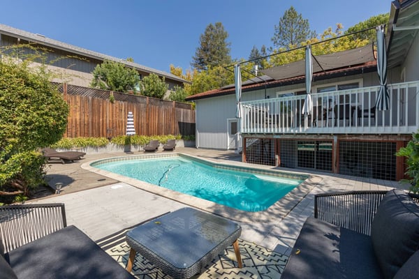 Backyard pool with lounge chairs, modern house with white railing deck, pergola, and pine trees