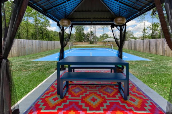 Covered pavilion with ping pong table overlooking blue tennis court in fenced backyard