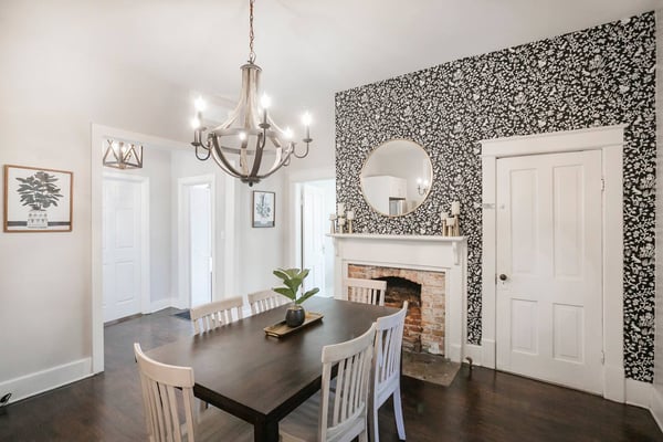 Dining room with black table, white chairs, chandelier, fireplace with round mirror, and black floral wallpaper accent wall