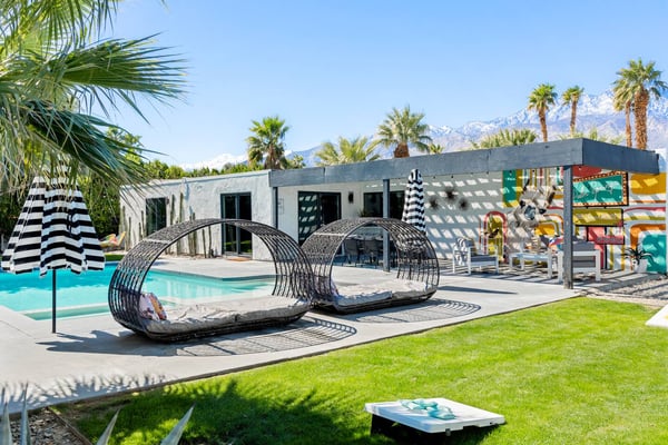 Modern poolside patio with turquoise pool, striped umbrella, woven lounge pods, white building, palm trees, and snow-capped mountains in background