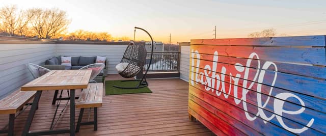 Rooftop deck at sunset with wooden dining table, hanging chair, seating area, and colorful painted fence at dusk.