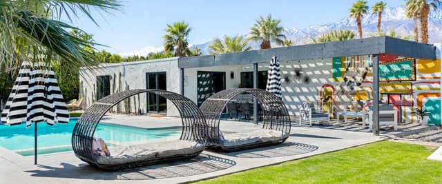Modern poolside patio with turquoise pool, striped umbrella, woven lounge pods, and mountain views with palm trees