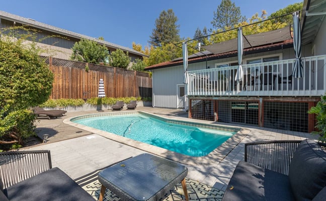 Backyard pool with lounge chairs, modern house with white railing deck, pergola, and pine trees