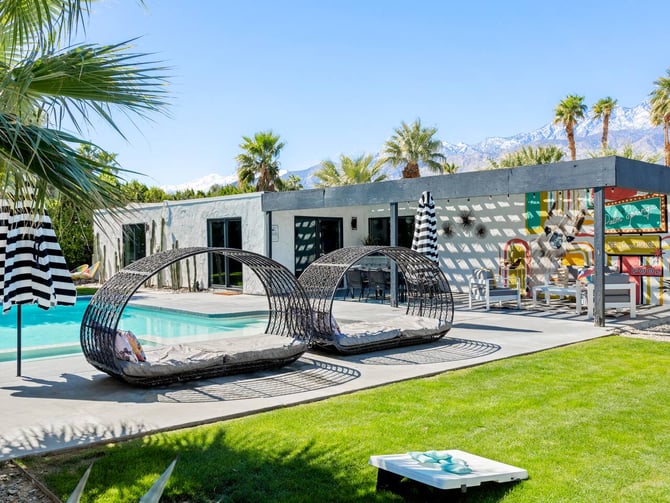 Modern poolside patio with turquoise pool, striped umbrella, woven lounge pods, white building, palm trees, and snow-capped mountains in background