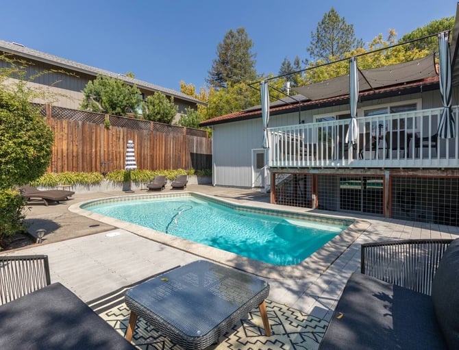Backyard pool with lounge chairs, modern house with white railing deck, pergola, and pine trees
